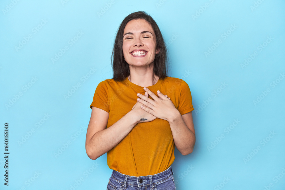 Young caucasian woman on blue backdrop laughing keeping hands on heart, concept of happiness. Young caucasian woman on blue backdrop laughing keeping hands on heart, concept of happiness.