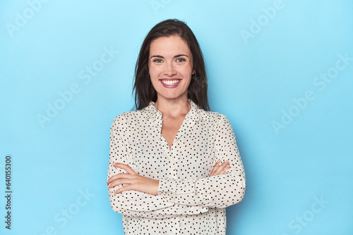 Young caucasian woman on blue backdrop who feels confident, crossing arms with determination.