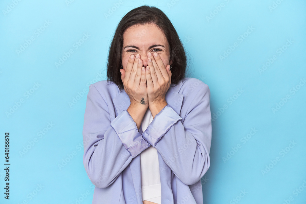 Woman in blue blazer on blue background laughing about something, covering mouth with hands. Woman in blue blazer on blue background laughing about something, covering mouth with hands.