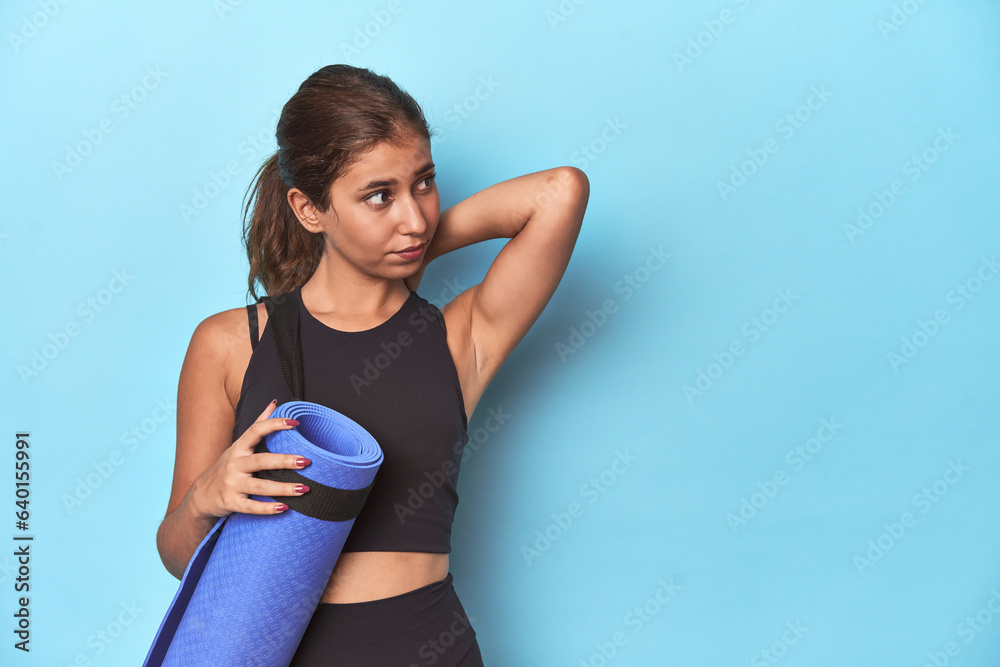 Young sportswoman with yoga mat, embracing wellness touching back of head, thinking and making a choice.