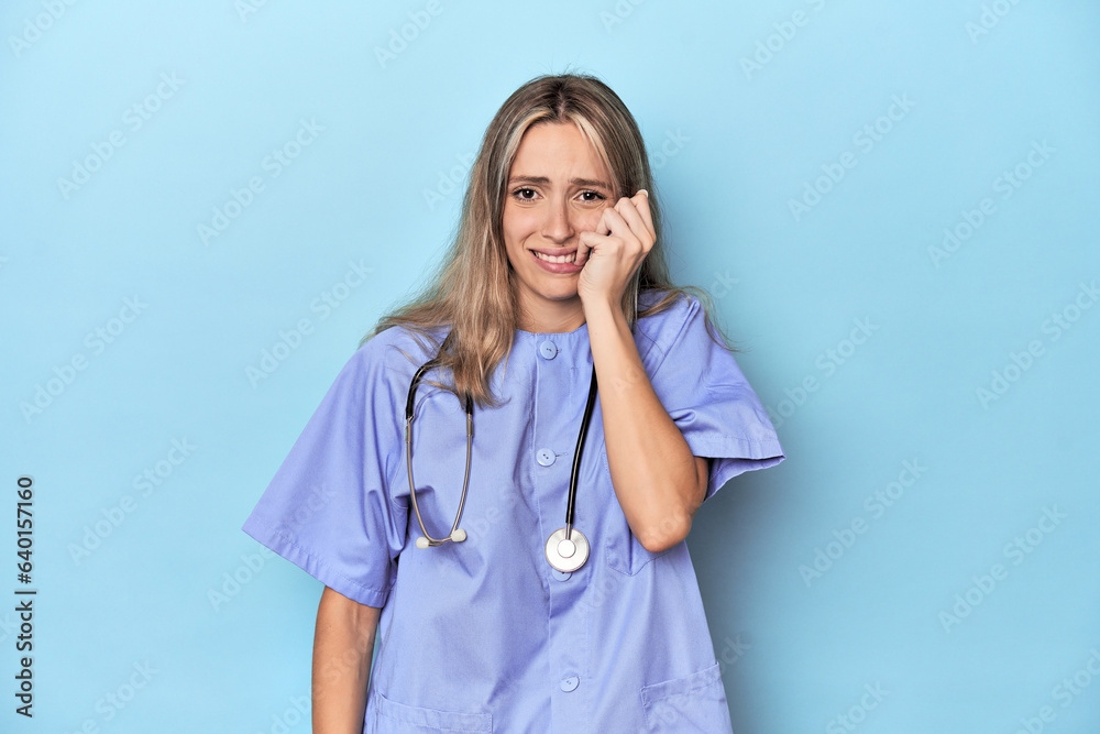 Young caucasian nurse in blue studio biting fingernails, nervous and very anxious. Young caucasian nurse in blue studio biting fingernails, nervous and very anxious.