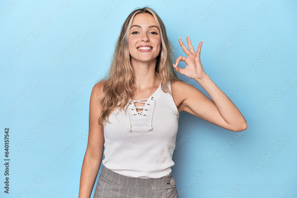Fototapeta premium Blonde young caucasian woman in blue studio cheerful and confident showing ok gesture.