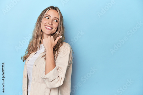 Fotografía Blonde young caucasian woman in blue studio points with thumb finger away, laughing and carefree