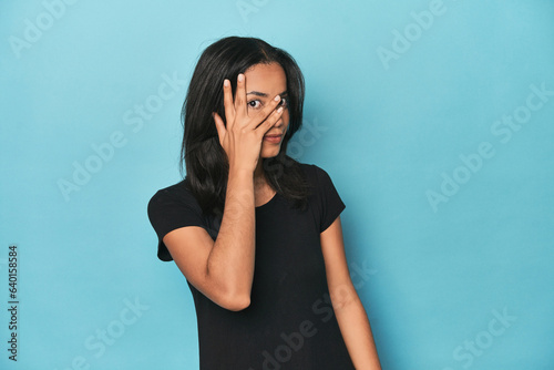Filipina young woman on blue studio blink at the camera through fingers, embarrassed covering face.