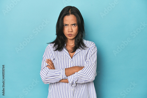 Filipina young woman on blue studio frowning face in displeasure, keeps arms folded.