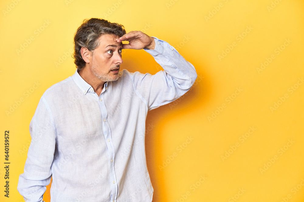 Middle-aged man posing on a yellow backdrop looking far away keeping hand on forehead. Middle-aged man posing on a yellow backdrop looking far away keeping hand on forehead.