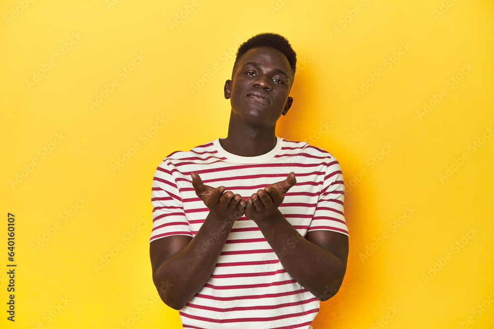 Stylish young African man on vibrant yellow studio background, holding something with palms, offering to camera. Stylish young African man on vibrant yellow studio background, holding something with palms, offering to camera.