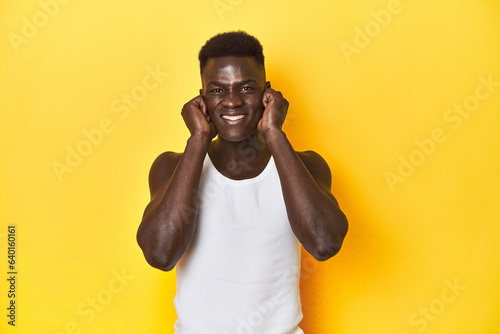 Stylish young African man on vibrant yellow studio background, covering ears with hands.