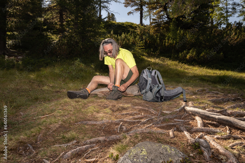 An adult gray-haired man with a backpack sits at the roots of a tree and ties a shoelace in the evening light