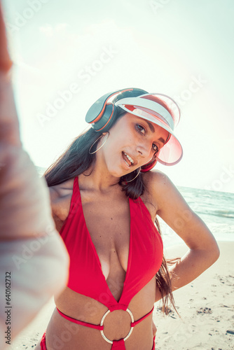 Photo shoot of a beautiful young woman posing wearing red sea costume, visor and headphones listeting to music during holidays with sky as background in a sunny day. Selfie photo with a smart phone