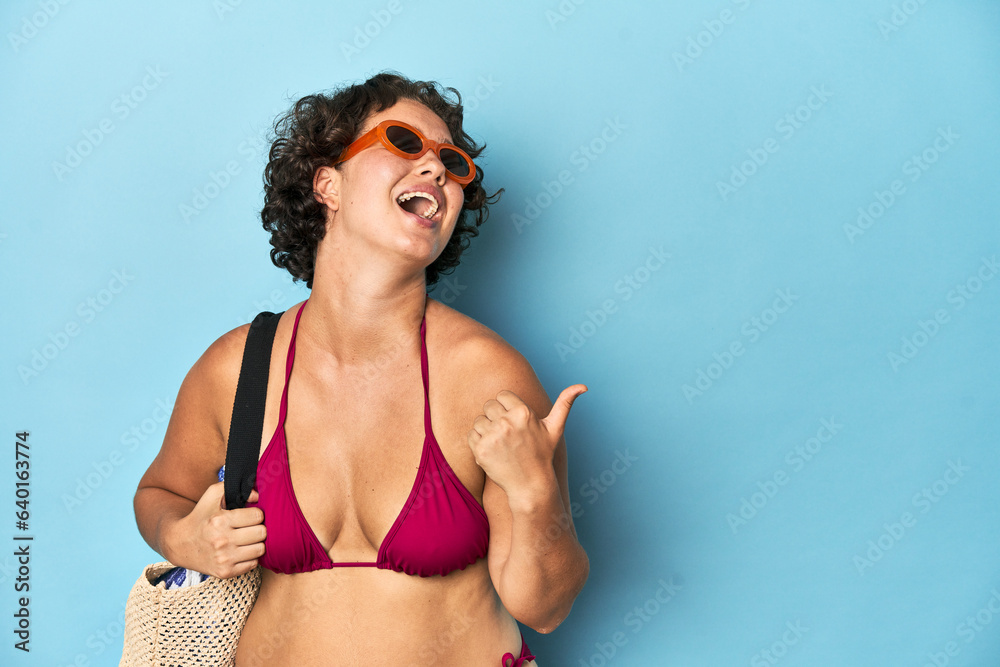 © Asier - Young woman in bikini with beach bag points with thumb finger away, laughing and carefree.