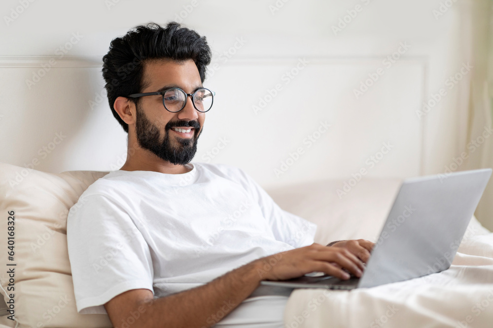 Smiling Young Indian Man Using Laptop While Sitting In Bed At Home Smiling Young Indian Man Using Laptop While Sitting In Bed At Home