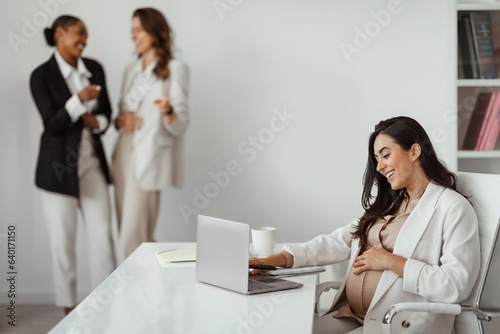 Businesswomen pointing at pregnant woman working on laptop in office, worker harassment and discrimination
