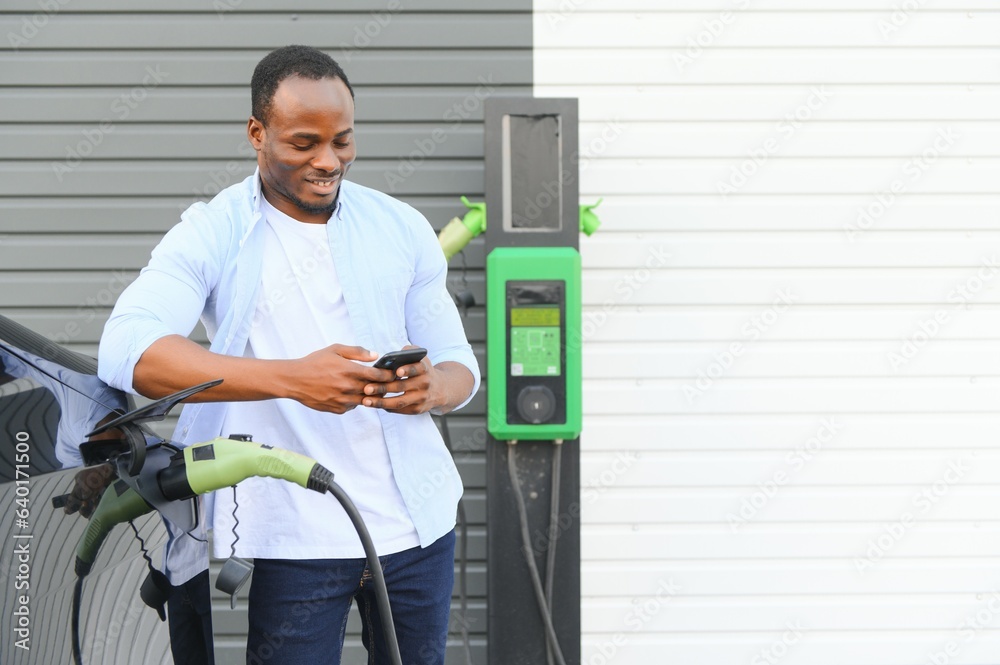 African man standing near electric car with charging cable in plug. Eco friendly vehicle charging on station. LIfestyles concept African man standing near electric car with charging cable in plug. Eco friendly vehicle charging on station. LIfestyles concept