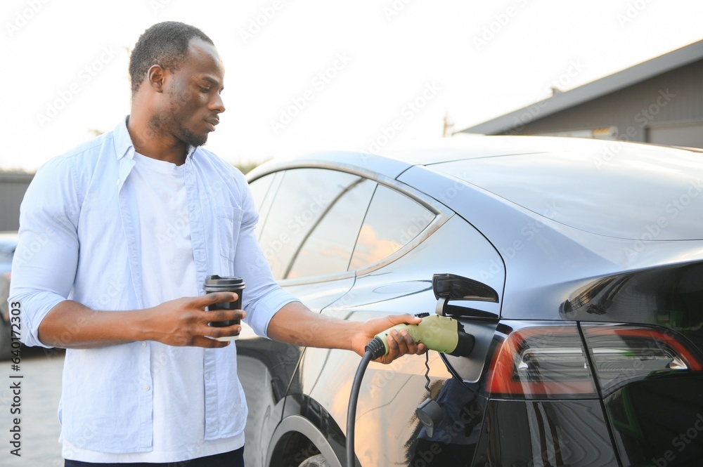 Serious african man holding charge cable in on hand, standing near luxury electric car.