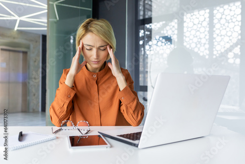 Tired woman at workplace having severe headache, depressed female worker holding hands on head, working inside office with laptop and paper work.