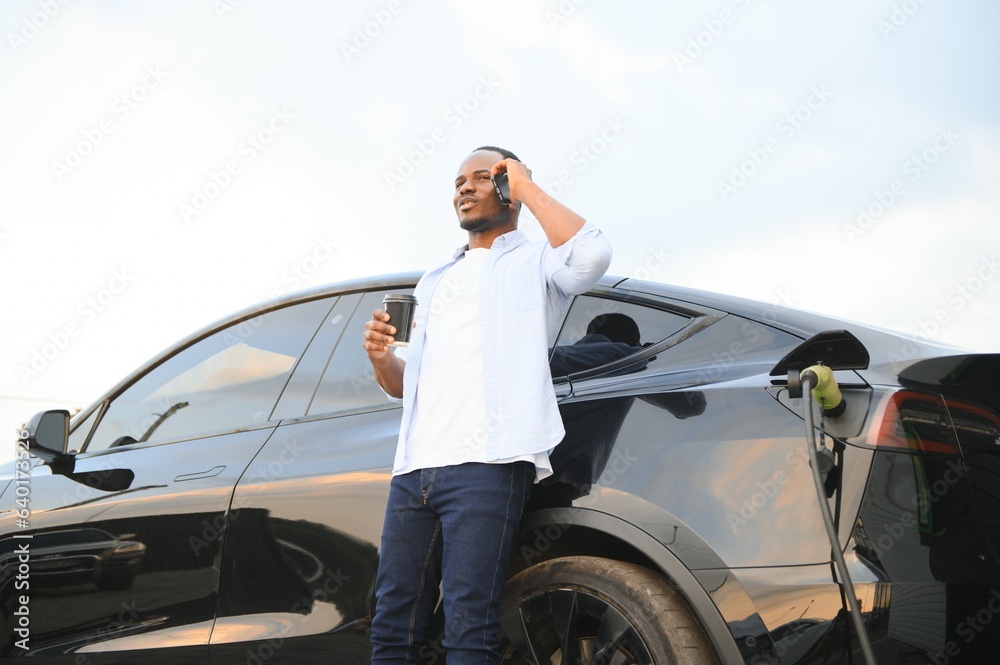 Happy young man with mobile phone charging car at electric vehicle charging station