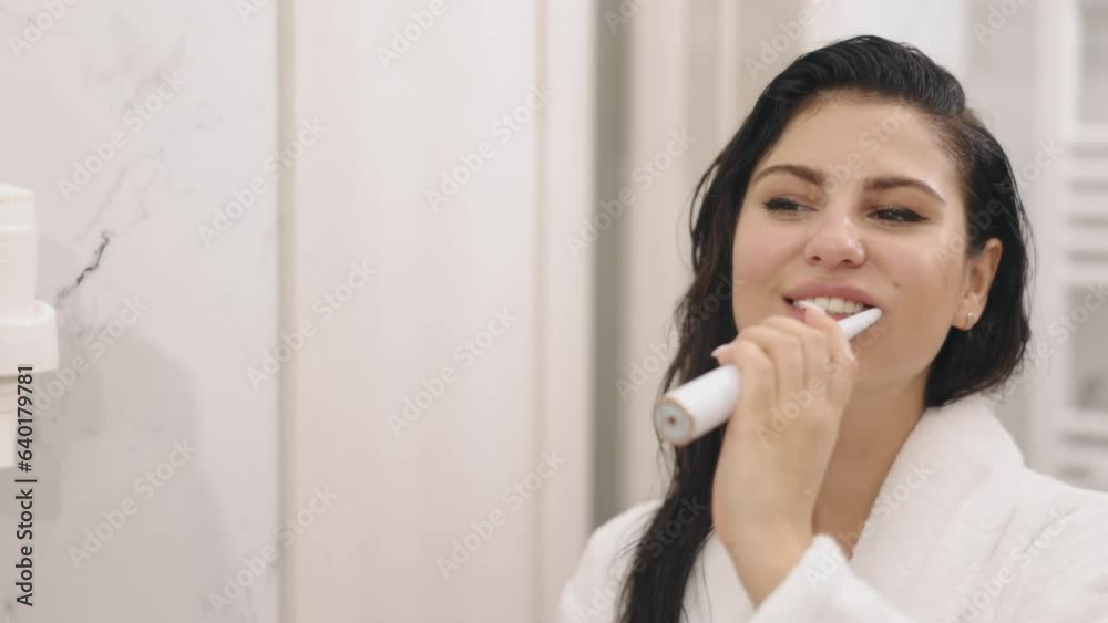 Adult Lady, Bathrobe Wearing, Dental Care. Lovely adult woman, fresh from shower, brushes teeth while checking reflection in bathroom mirror, wearing white, silky bathrobe Adult Lady, Bathrobe Wearing, Dental Care. Lovely adult woman, fresh from shower, brushes teeth while checking reflection in bathroom mirror, wearing white, silky bathrobe
