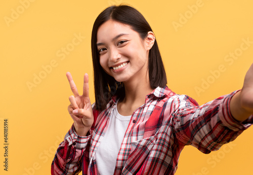 Glad japanese woman taking selfie and showing peace sign with hand posing isolated on yellow background, studio shot