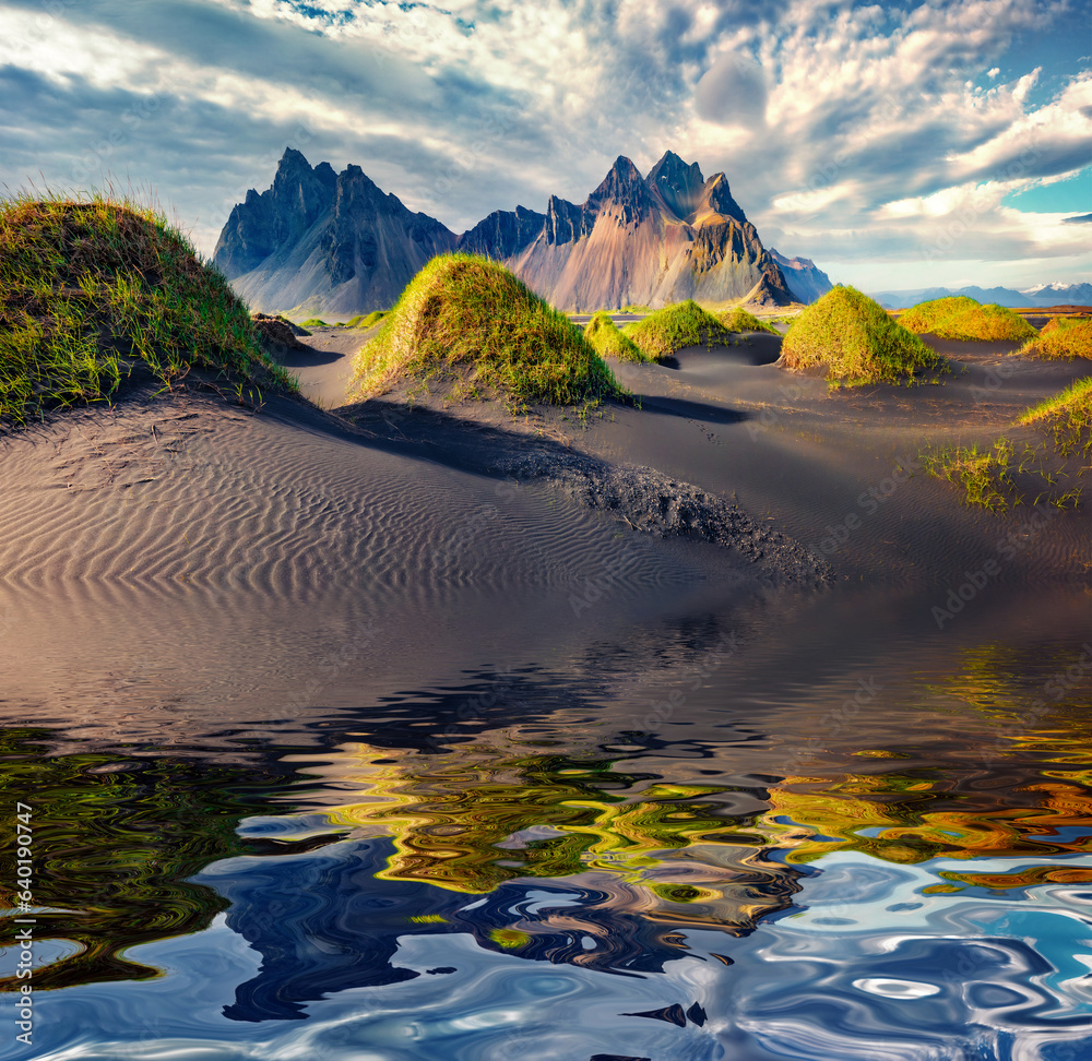 Fototapeta premium Stokksnes cape with Vestrahorn (Batman Mountain) reflected in the calm waters of Atlantic Ocean. Amazing summer landscape of black sand dunes with green grasson the top. Travel to Iceland.
