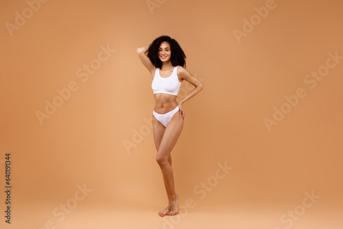 Slender latin lady with perfect body shape posing in white underwear, brown background, full length, copy space