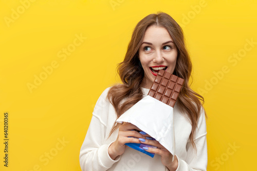 Fotografie young girl eats and bites big chocolate bar and thinks and dreams on yellow isol