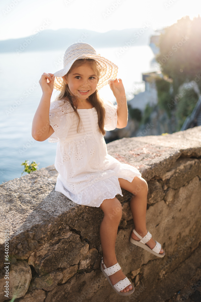 Cute little girl in beautiful white dress and hat sitting on bordure on heigh coast in front of sea and mountains Cute little girl in beautiful white dress and hat sitting on bordure on heigh coast in front of sea and mountains