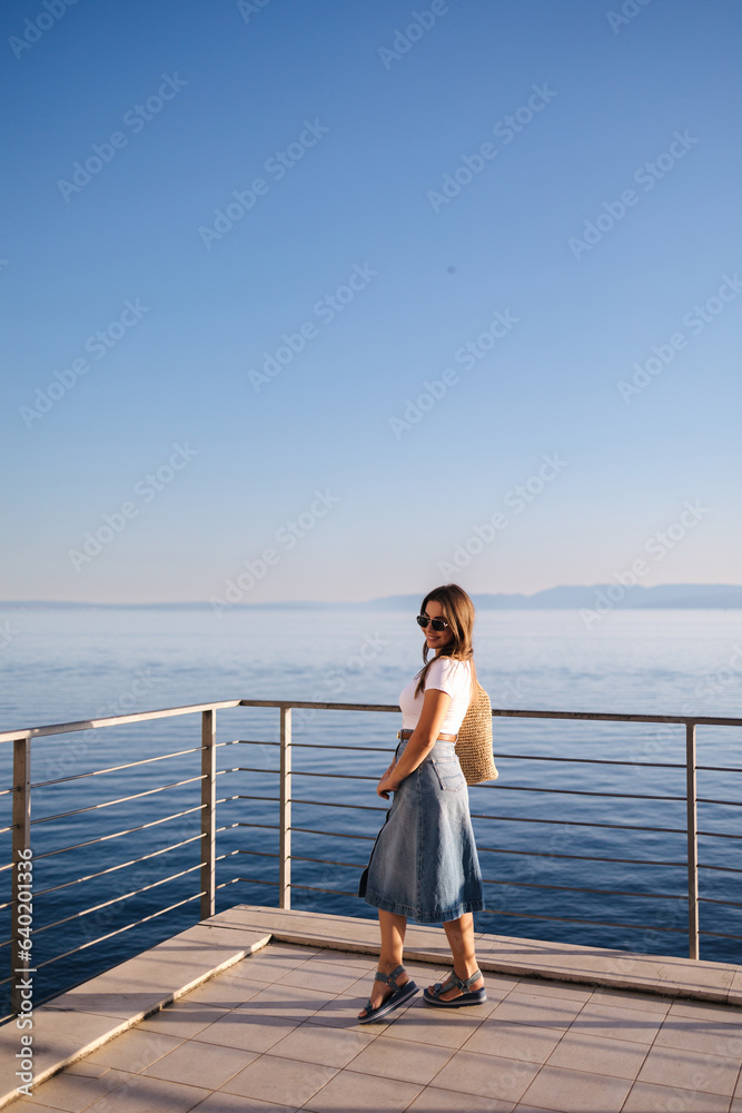 Beautiful woman stand on balcony an looking on sunset near the sea. Female in denim style with knitted bag
