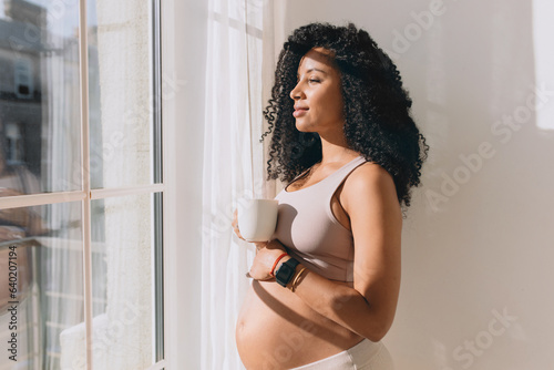 Charming lovely pregnant female in crop top patting her belly standing in front of window under sun rays, drinking coffee or tea in morning, talking too her baby, imagining their future life together