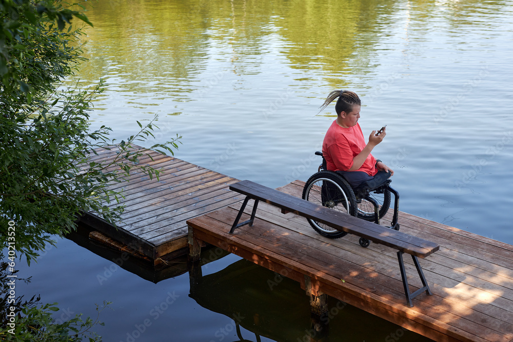 High angle portrait of young man with disability sitting in wheelchair by lake and using smartphone, copy space High angle portrait of young man with disability sitting in wheelchair by lake and using smartphone, copy space