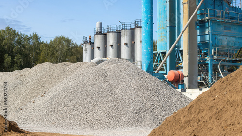 On the territory there is a pile of gravel and sand in close-up against the background of the production equipment of the asphalt concrete plant. Clear sunny summer day at the factory.