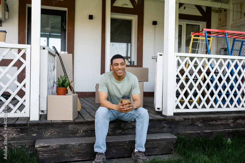 smiling african american man holding takeaway coffee while sitting on porch of new house