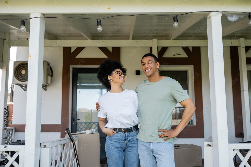 smiling african american couple hugging while standing on porch of new house