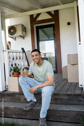 smiling african american man holding coffee to go near packages on porch of new house
