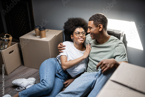 cheerful african american woman hugging boyfriend near coffee to go and boxes in new house