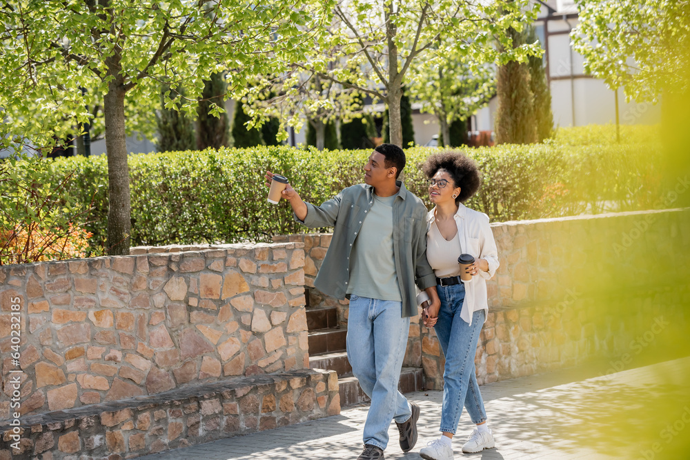 african american man with coffee to go pointing on urban street near cheerful girlfriend