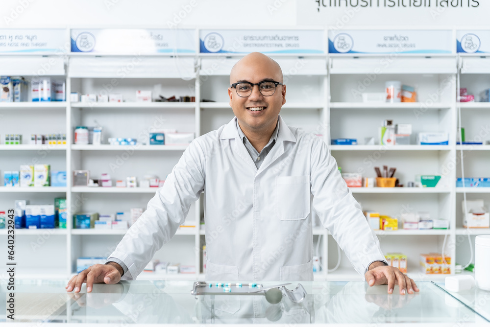 Happy handsome asian male pharmacist wearing eyeglasses and lab coat standing and looking at camera, He feels good, trustworthy and proud of his work in the pharmacy drugstore.	