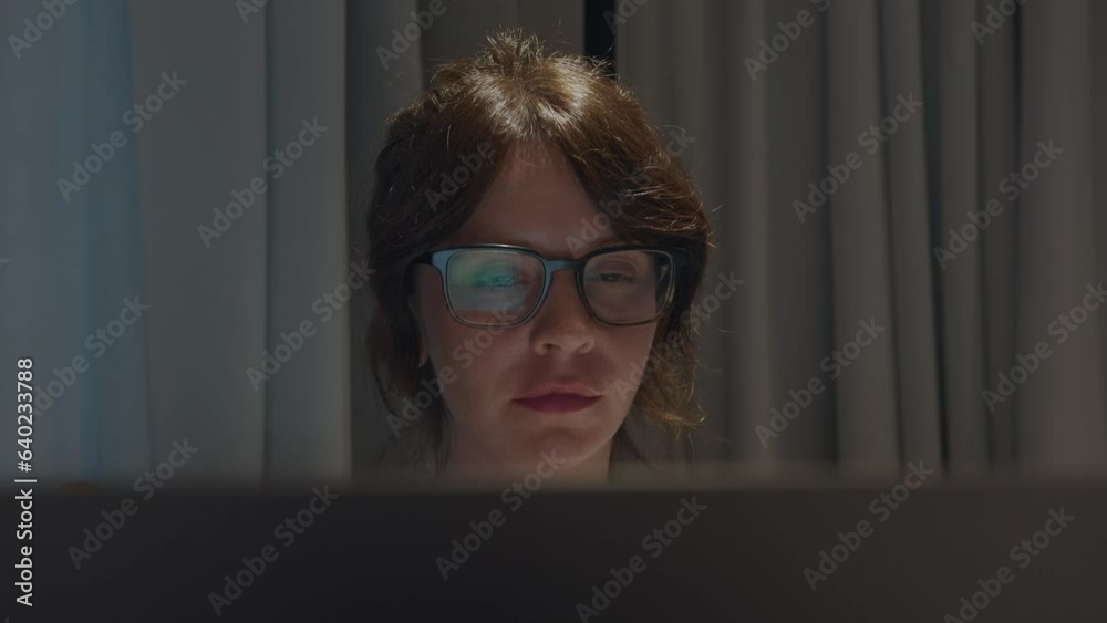 Concentrated young woman closeup portrait wearing eyeglasses, looking at the laptop screen. Pensive woman working at night from home, curtains on background Concentrated young woman closeup portrait wearing eyeglasses, looking at the laptop screen. Pensive woman working at night from home, curtains on background