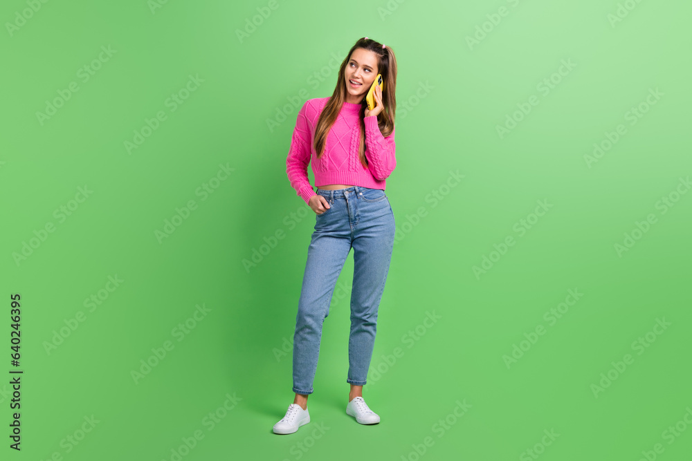 Full size photo of gorgeous lovely girl wear pink pullover jeans look empty space talk on smartphone isolated on green color background Full size photo of gorgeous lovely girl wear pink pullover jeans look empty space talk on smartphone isolated on green color background