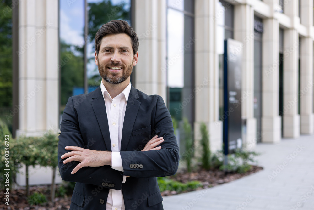 Portrait of a handsome young businessman, manager, leader standing in a suit near the office center and crossing his arms confidently smiling at the camera Portrait of a handsome young businessman, manager, leader standing in a suit near the office center and crossing his arms confidently smiling at the camera