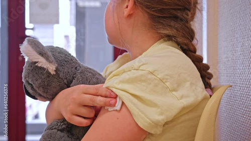 Caucasian Girl Kid Hugging Teddy Bear Sitting in Clinic Waiting Room After Vaccine Shot Pressing Gauze Pad Against Needle Injection to Stop Bleeding  photo