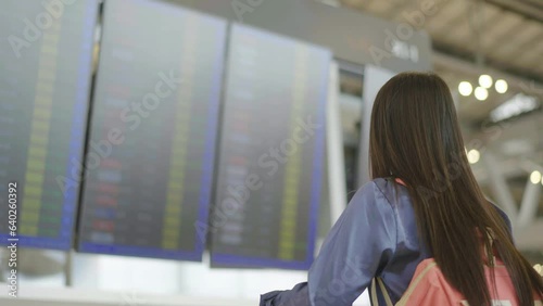 Young asian woman with passport and boarding pass as a hand stand in front of the airline time schedule board looking at the flight information board at the airport terminal, checking her flight