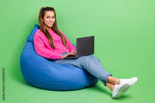 Full length photo of clever woman with tails dressed pink sweater sit on pouf with laptop typing email isolated on green color background
