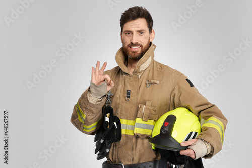 Happy dark-haired firefighter in uniform gesturing okay sign with hand in studio. Portrait of smiling bearded fireman showing ok, while looking camera, isolated on gray. Concept of work, hand gesture.