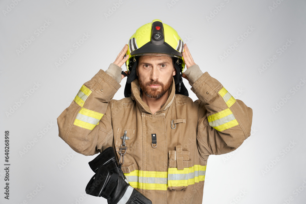 Focused caucasian firefighter putting on yellow protective helmet before working shift. Portrait of serious man wearing fireman's uniform, looking at camera, isolated on gray. Work, safety concept. Focused caucasian firefighter putting on yellow protective helmet before working shift. Portrait of serious man wearing fireman's uniform, looking at camera, isolated on gray. Work, safety concept.