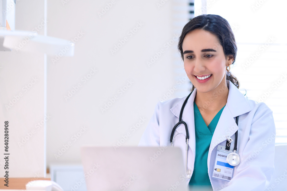 Smiling professional female doctor in white medical coat working with laptop computer, sitting at the table near the window in hospital office, looking at laptop screen. Smiling professional female doctor in white medical coat working with laptop computer, sitting at the table near the window in hospital office, looking at laptop screen.