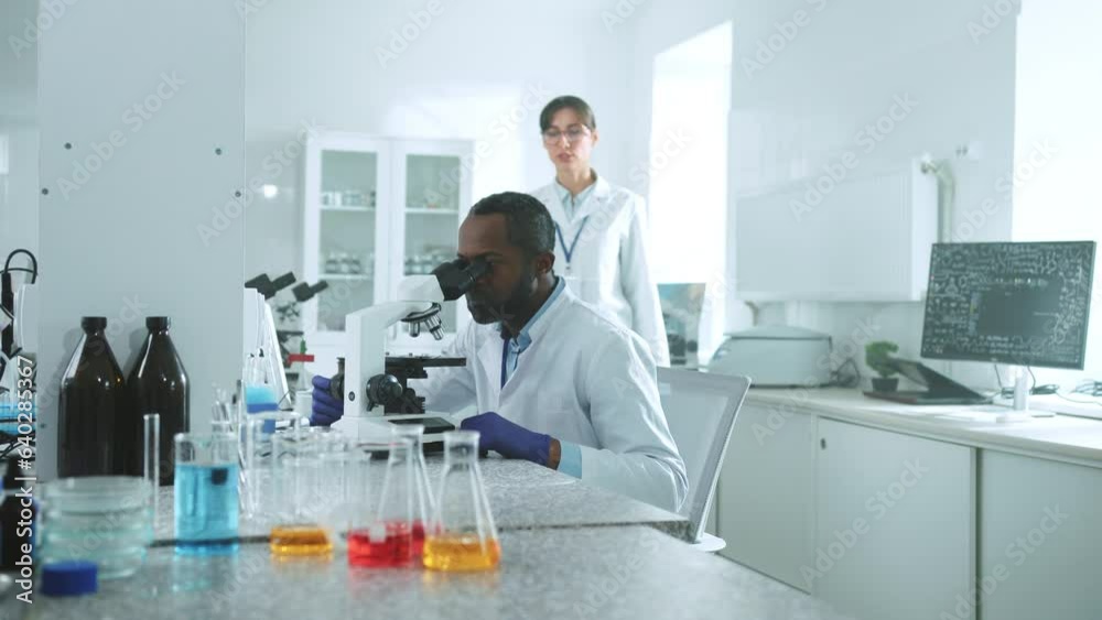Two scientists in uniform looking under a microscope talking discuss wonder in the laboratory. African American man and a Caucasian woman. Research, biochemistry, pharmaceutical medicine Two scientists in uniform looking under a microscope talking discuss wonder in the laboratory. African American man and a Caucasian woman. Research, biochemistry, pharmaceutical medicine