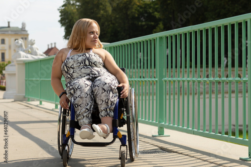 Beautiful Young Woman With Short Height Walks Over Bridge In Wheelchair In City at Summer Day. Female Adult With Disability. Copy Space For Text. Horizontal Plane