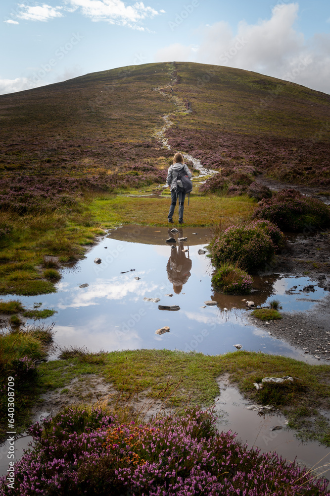 A young woman walking towards peak in Irish mountains. Water puddle with reflection in foreground. A young woman walking towards peak in Irish mountains. Water puddle with reflection in foreground.