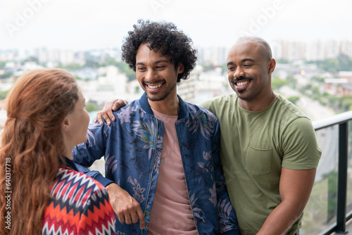 Same-sex couple talk to friend and smile on apartment balcony